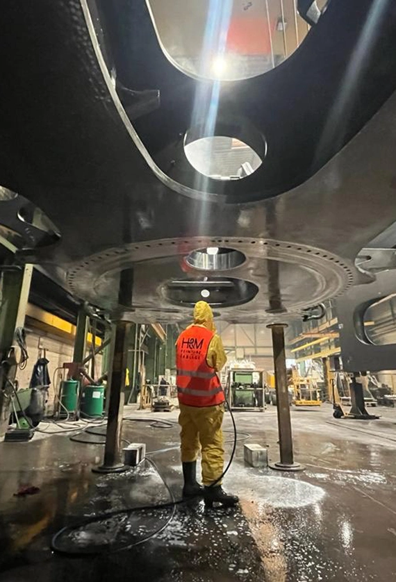 Dans cette image, des professionnels réalisent un décapage méticuleux d'une surface à l'aide d'un abrasif projeté à haute pression depuis une cabine spécialisée. On peut également observer une zone préparée pour la métallisation, juste à côté d'une section où la peinture vient d'être appliquée. L'expertise et la précision des professionnels sont manifestes dans chaque détail du processus